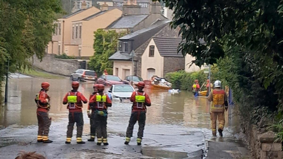 A group of firefighters stand in front of a flooded road. A white car is submerged in the water.