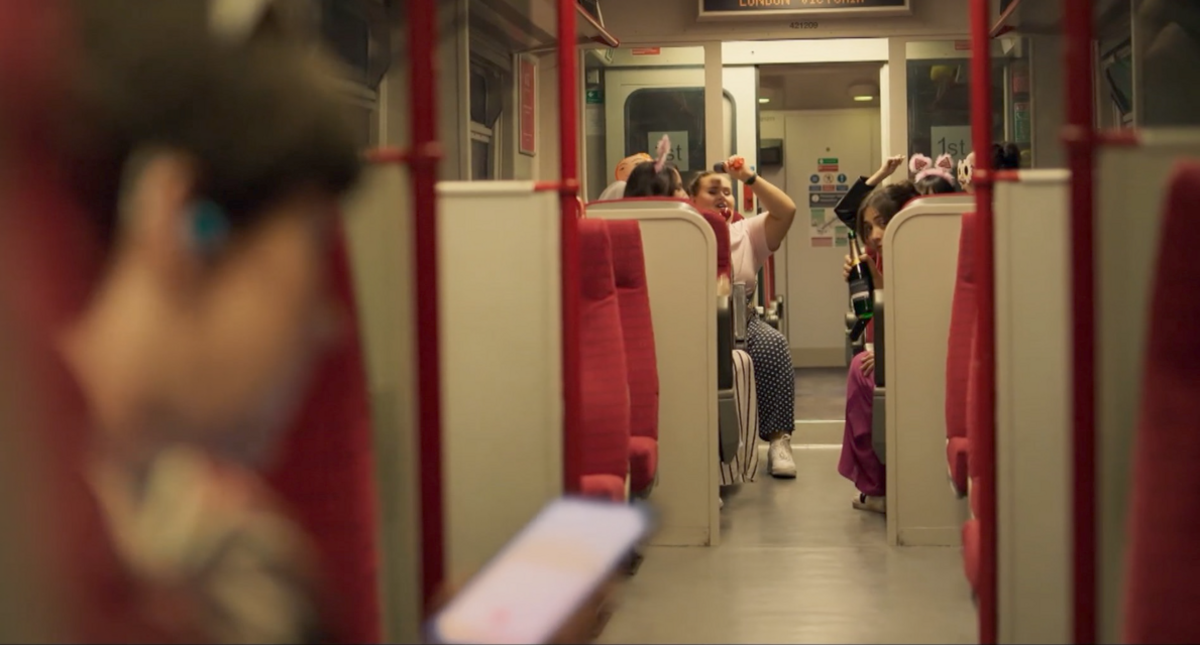 A group of passengers can be seen at one end of a train carriage. In the foreground a woman looks at a mobile phone screen. She is out of focus.