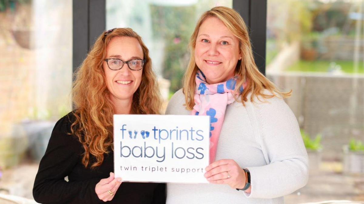 Sharon Darke with long curly hair, wearing a black top, and Suzie Scofield with long straight blonde hair wearing a white top. They are holding a sign which says "Footprints baby loss, twin triplet support"