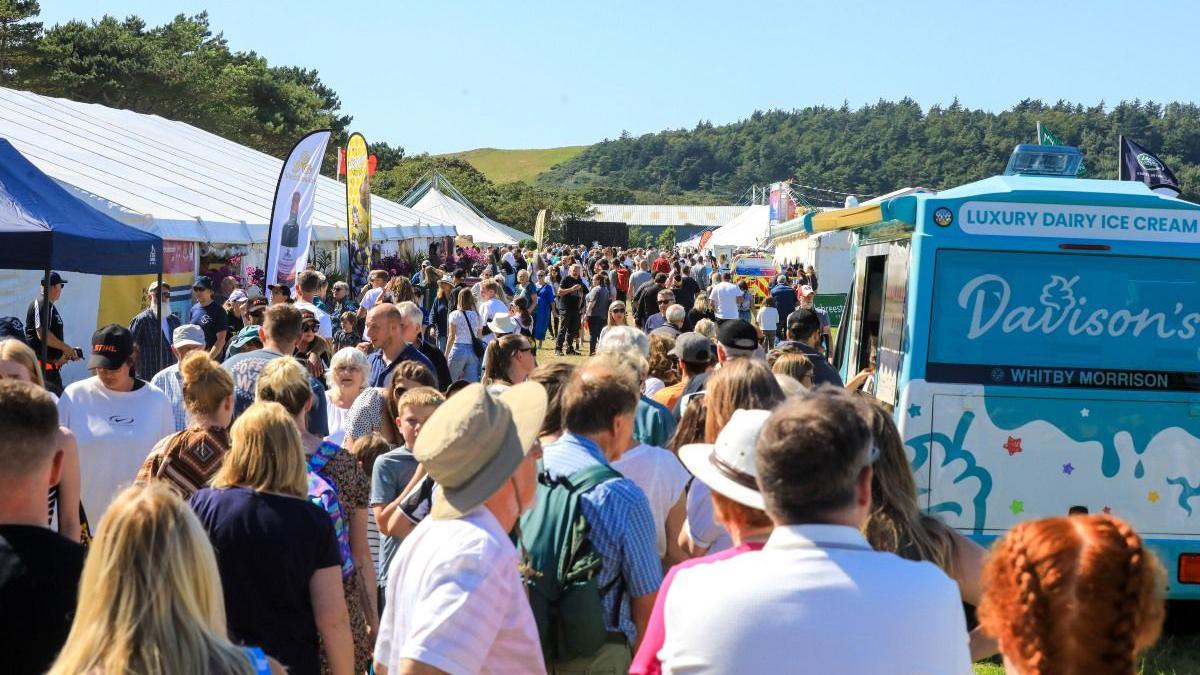 Crowds of people in summer clothes among the marquees and an ice cream van on the show field.