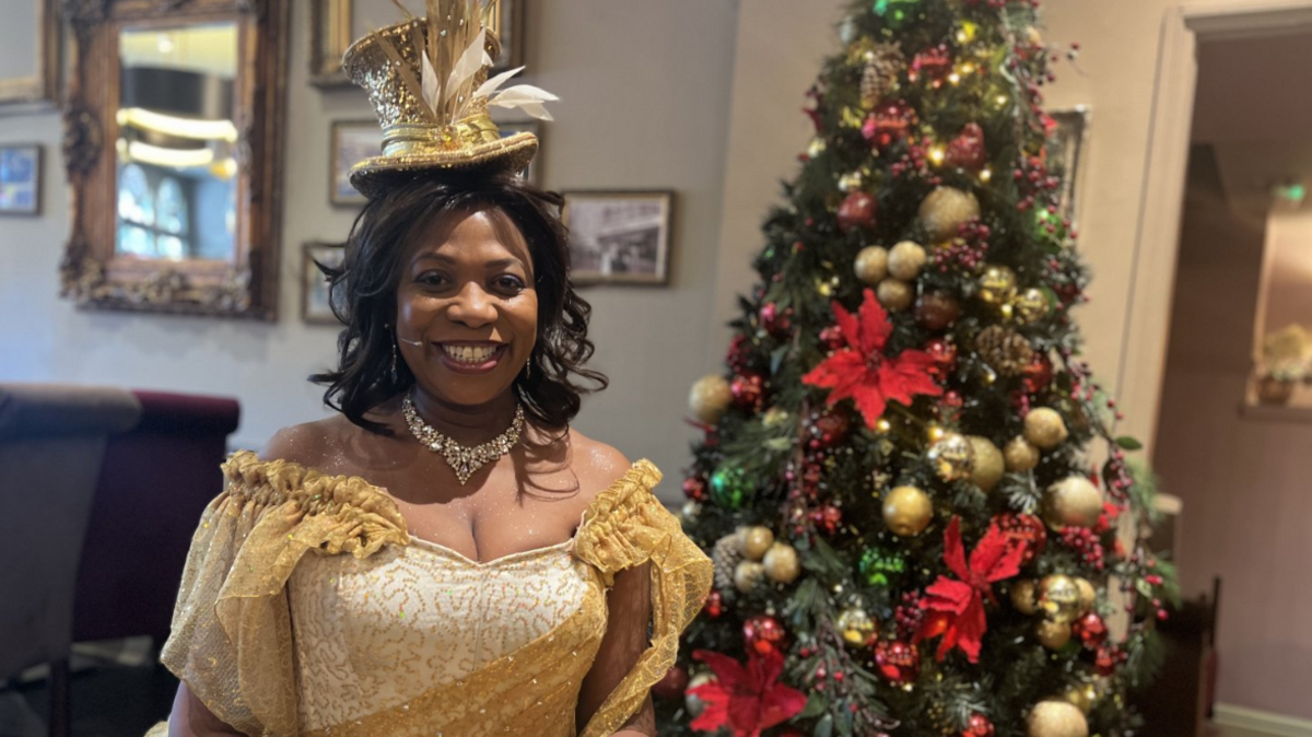 Brenda Edwards wearing her Candy Floss dress which is gold and glittery with big sleeves. She also has a diamante necklace on and a gold top hat over her brown curly hair. There is a Christmas tree behind her to the right.