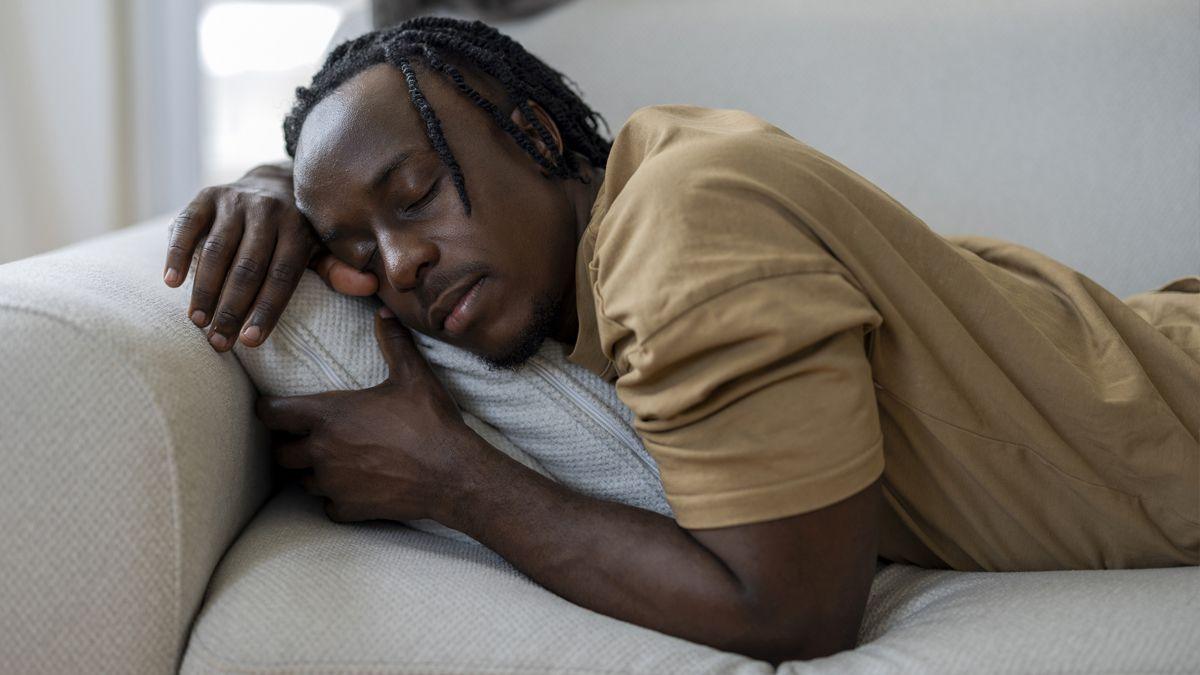 A man sleeping on a sofa at home with a grey cat resting in the background. His relaxed posture and closed eyes suggest a peaceful nap, rest, fatigue, or quiet leisure time