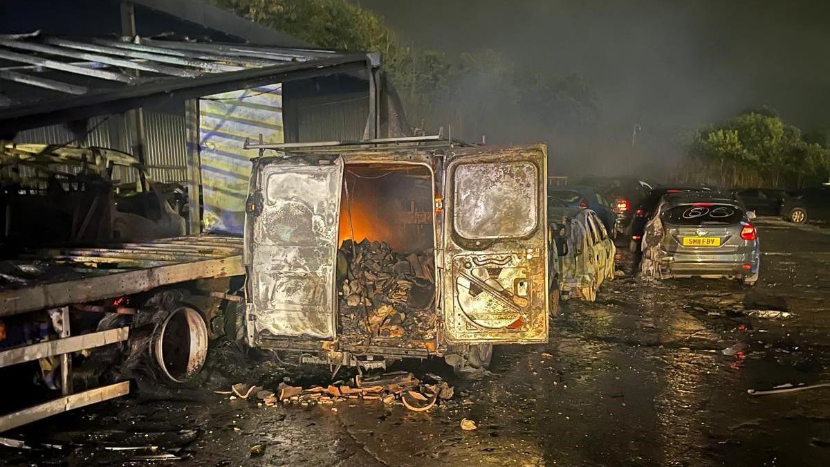 A burned out van full of scrap metal has an orange glow emanating from inside. It is night time on on an industrial estate surrounded by other fire debris.