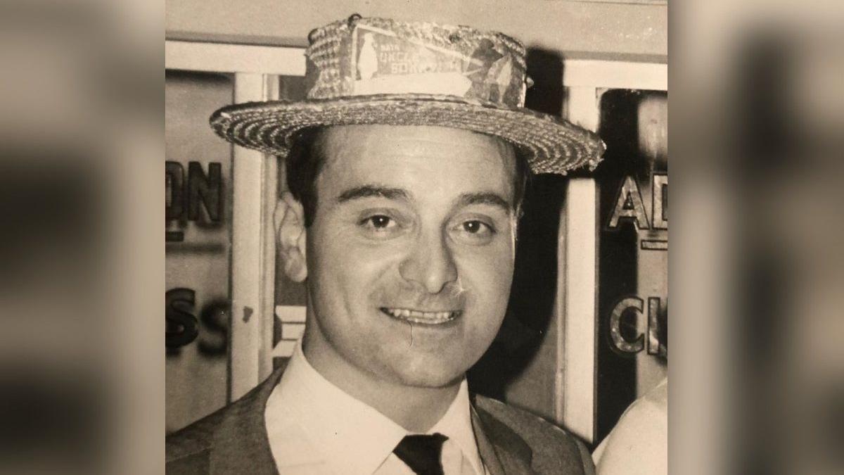 Black and white image of a man wearing a straw hat, suit, shirt and tie. He is smiling. 