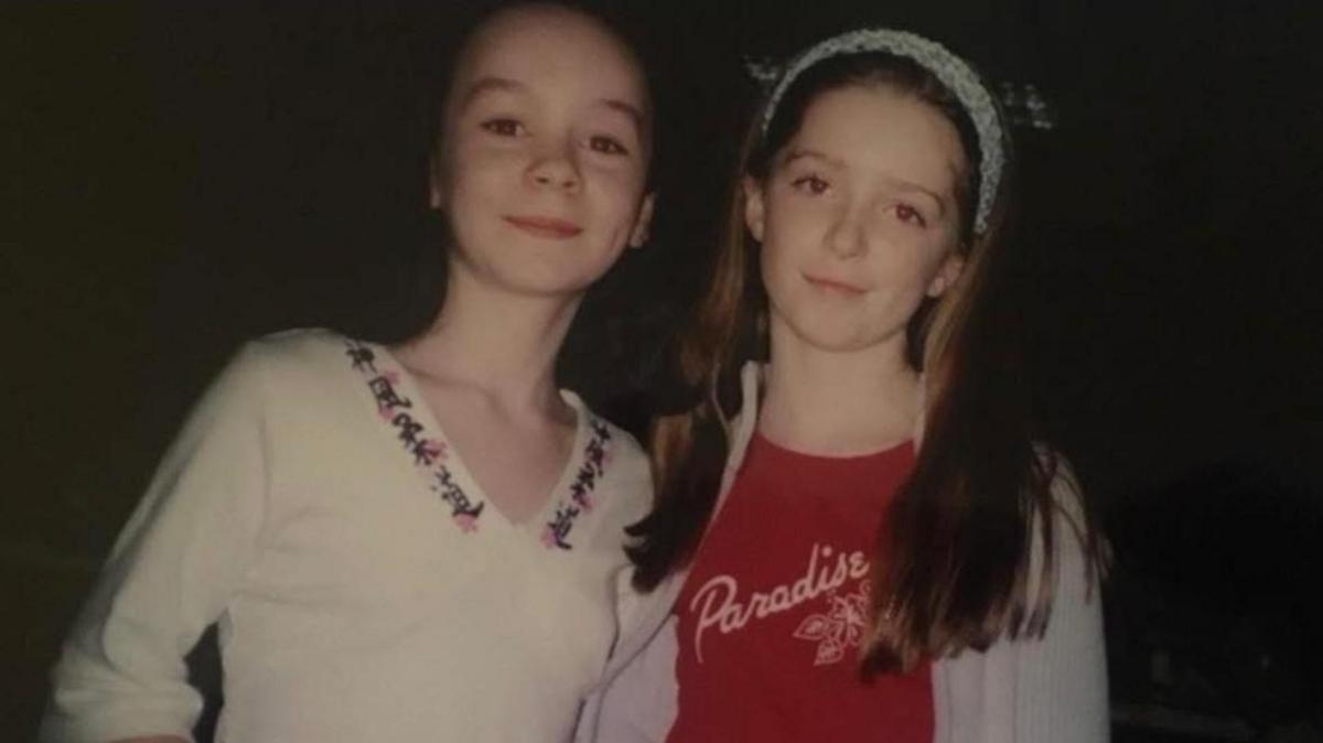 Two young girls stand together smiling at the camera. They both have brown hair. The girl on the left wears a white top with a floral pattern at the collar, while the other girl wears a red t-shirt and blue headband.