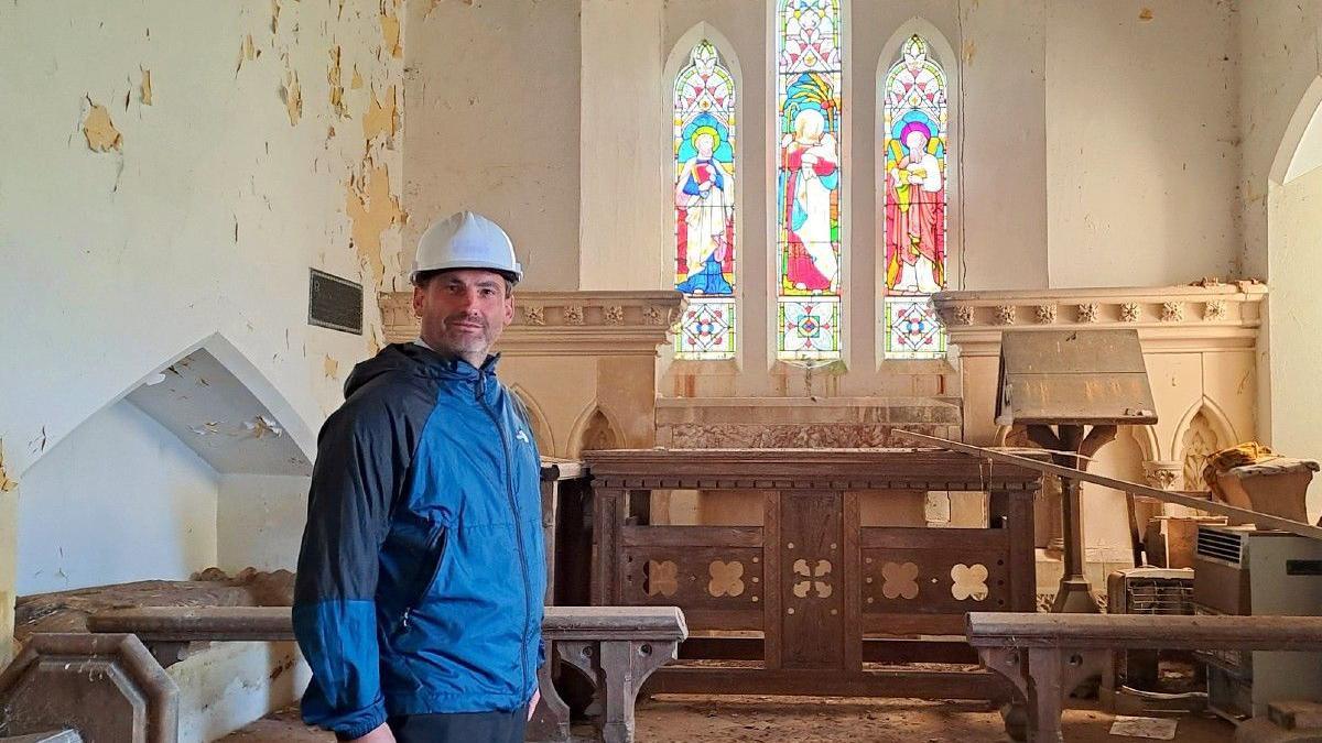 Sam Pratley stands in the chancel, which is in disrepair and the paint is crumbling from the walls. There are three stained glass windows and there is a stone sculpture behind him.