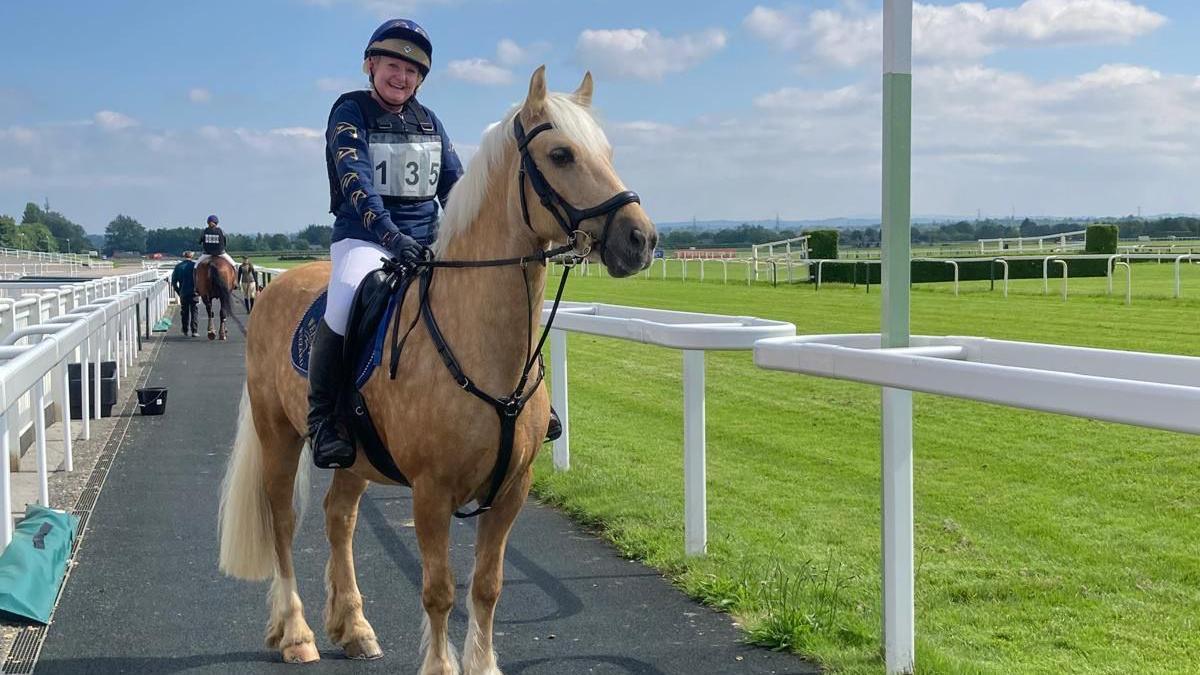 Karen Tidman sits on her horse and smiling. She is wearing dark blue silks, bearing the number 135, and white jodhpurs. She and her horse are on a tarmac path next to a racecourse.