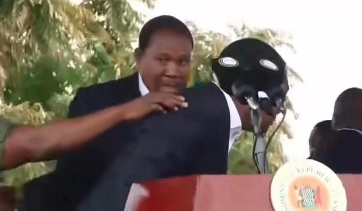 President Hakainde Hichilema crouches behind a microphone. A hand can be seen reaching out from the left hand side of the frame touching the president on the back. A man behind the president is looking nervous.