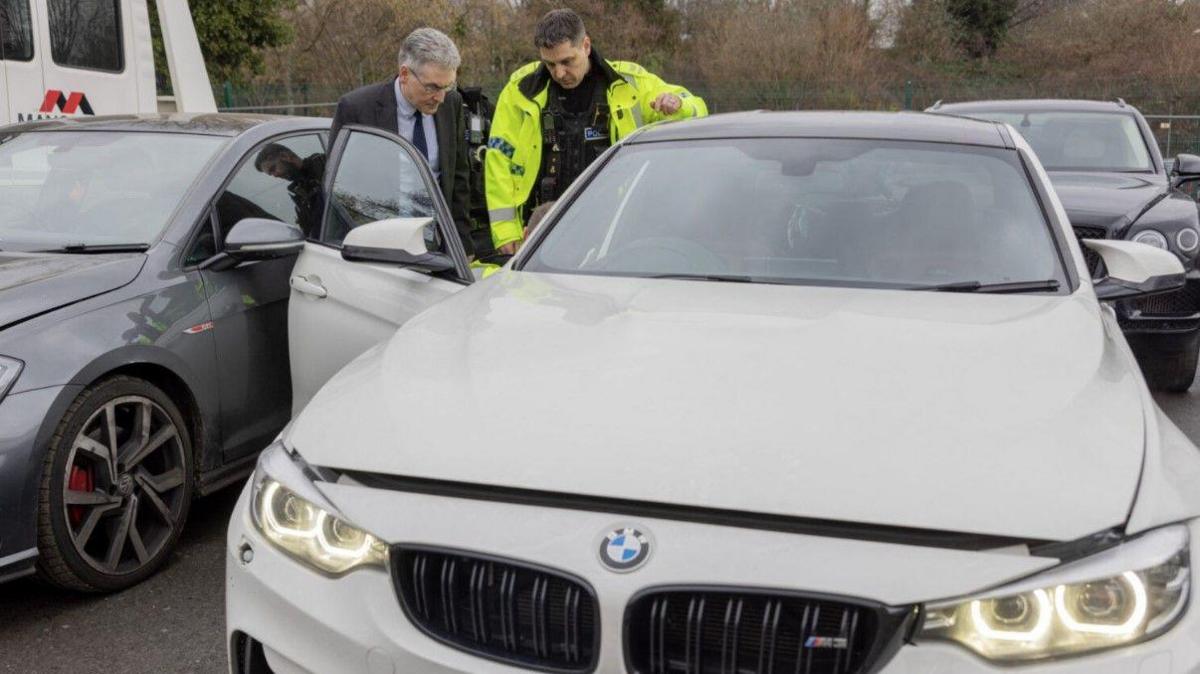 A white BMW, pictured by the bonnet, with its front lights on. Simon Foster is standing up, next to another man, looking inside it. Other vehicles are in the background.