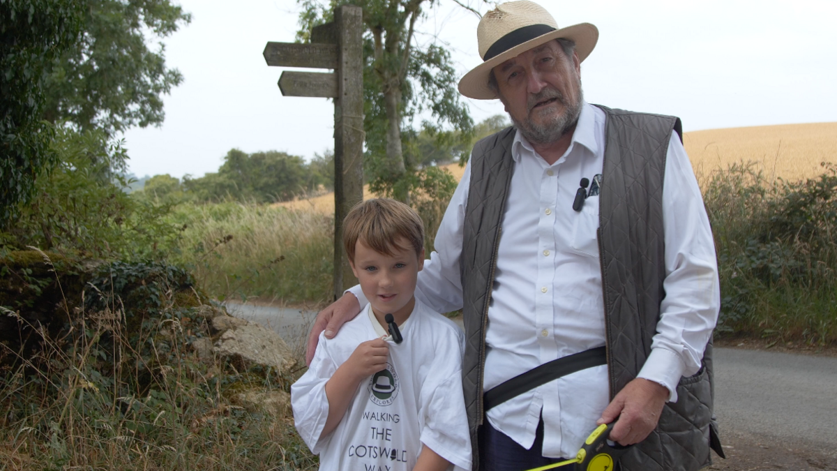 Mr Shuckburgh speaking to the camera on his walk. He has his arm around his grandson Huxley. The boy has a microphone clipped on his T-shirt. The T-shirt is reading The Cotswold Explorer walking the Cotswold Way.