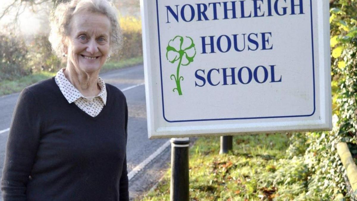 Viv Morgan standing next to a Northleigh House School sign. Behind her is a road and the sign is on a grass bank. 