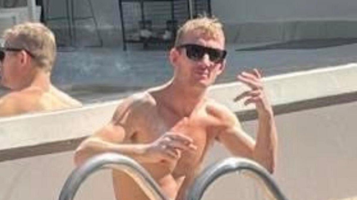 Craig Yorke. He is sitting by a pool without a shirt on and is wearing sunglasses. He is slim but muscular and is waving at the camera.