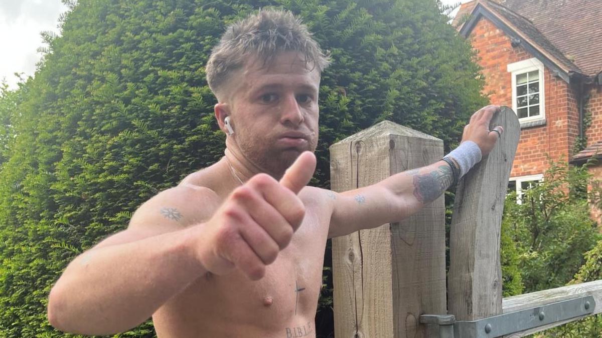 Tommy, a young man, has his top off and is doing a thumbs up at the camera with a tired look on his face. He is resting his other hand on a wooden gate by a house. He has several tattoos, including a cross on his sternum. 