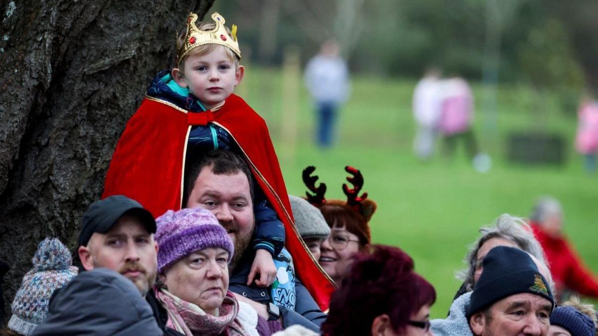 A boy wearing a plastic gold crown and red cape sits on the shoulders of a man with a crowd of people gathering ahead of the Christmas Day morning church service attended by the royal family at St Mary Magdalene Church in Sandringham, Norfolk. People are wearing wool hats and and chat among themselves.