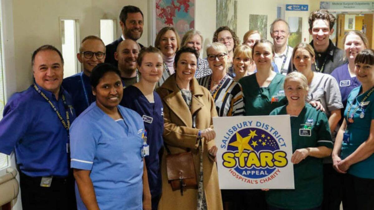 The actress Olivia Colman holds a plaque devoted to the Stars Appeal charity. She is surrounded by staff and volunteers at Salisbury Hospital. They are standing tightly together, huddled around the actress as they all smile at the camera. 