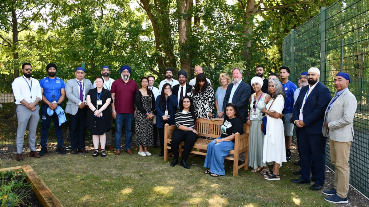 Dozens of people stand in a line around a memorial bench in a garden and smile. Two women sit on the brown bench. Trees are behind them.