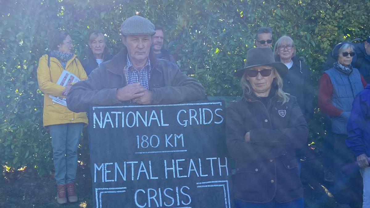 Paul and Jackie Thompson are facing the camera with expressionless faces. They are standing with a black sign that reads, in white writing, 'National Grids 180km mental health crisis'. Other protesters can be seen in the background.