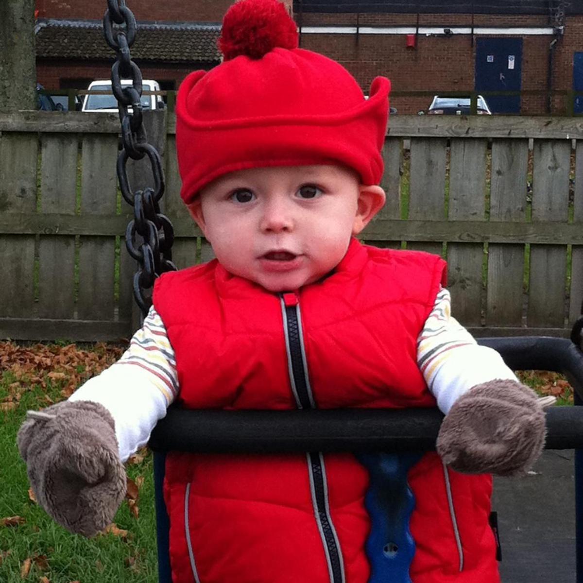 Baby Gethin is wearing a red hat and coat and is sat in a swing.