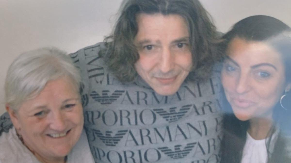 A middle-aged man with long dark hair stands with has arms around a younger and an older woman.