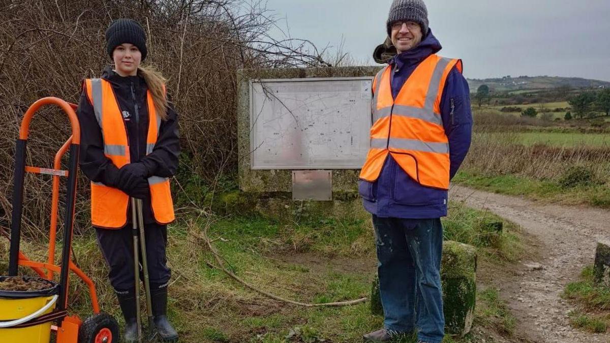 Wearing a high vis jacket, gloves and boots Elsa holds litter-picking tools and stands in front of a bucket on a trolley. She is accompanied by her father who is wearing similar protective gear.