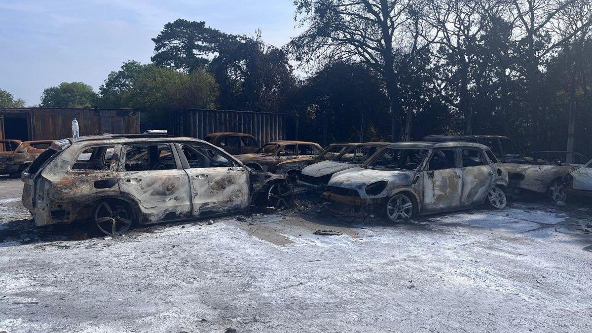 The charred remains of cars stand in a yard covered in ash.