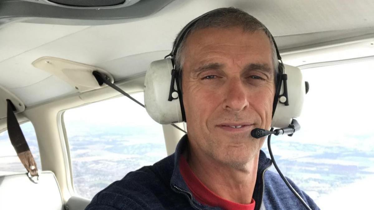 A smiling middle-aged man in the seat of a small plane, flying above fields. He is earing a headset.