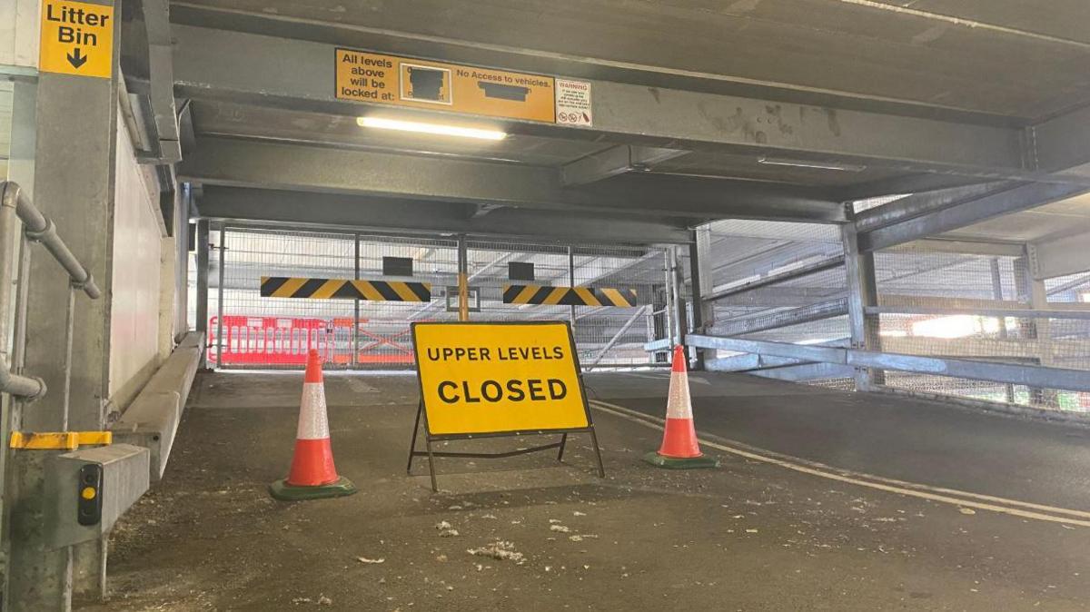 A upper level closed sign sits in the middle of a ramp leading up to further floors in a car park. Bollards are there too.