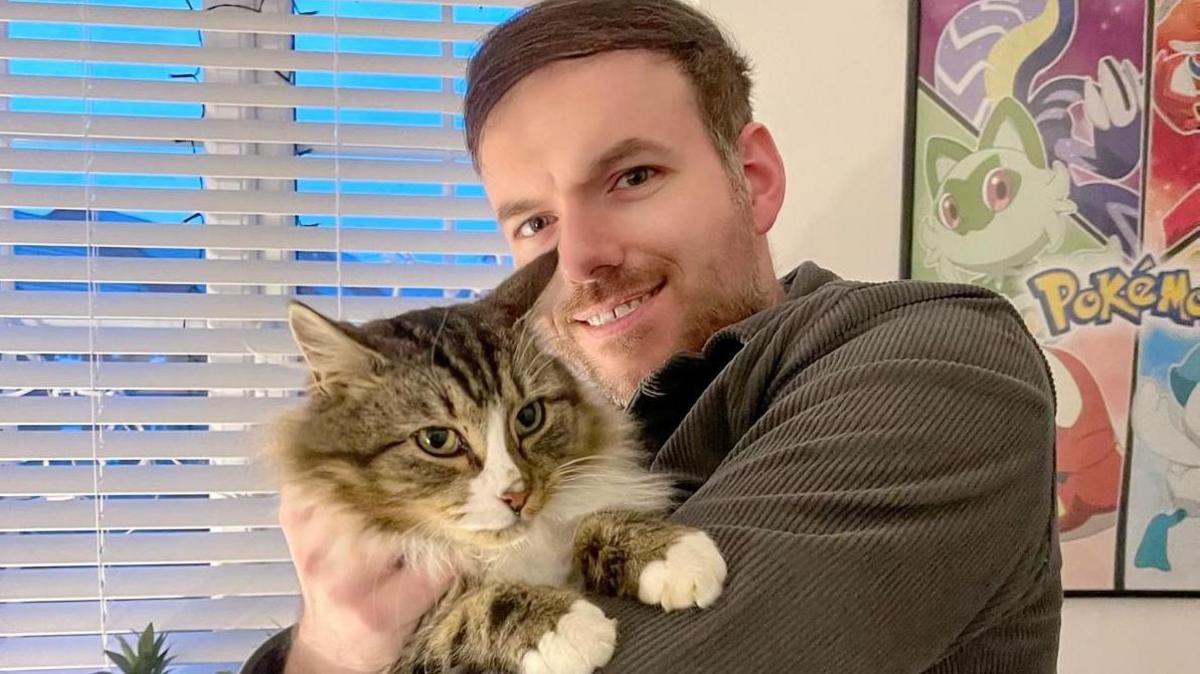 Jonathon Marlow with brown hair and beard and brown shirt holding tabby cat Rhubard in his arms smiling