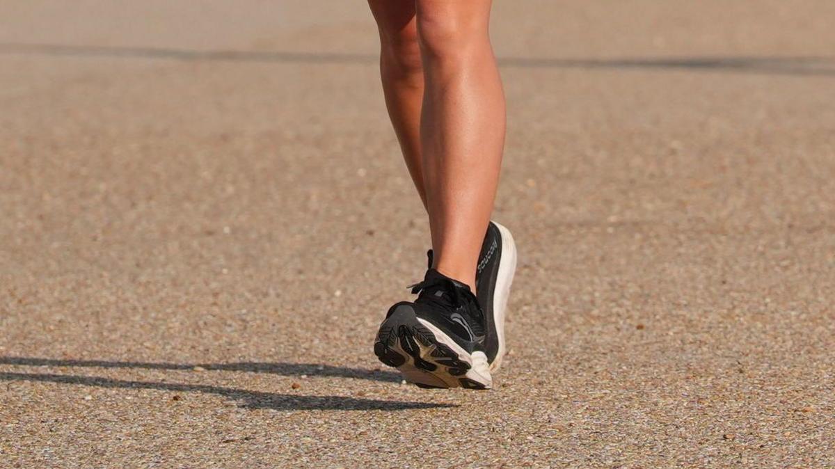 A close-up image of legs and black and white trainers running. The ground is a sandy colour.