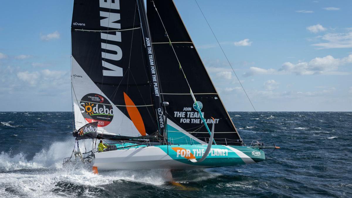 A racing yacht with a white and blue hull and black and white sails going fast with spray behind.