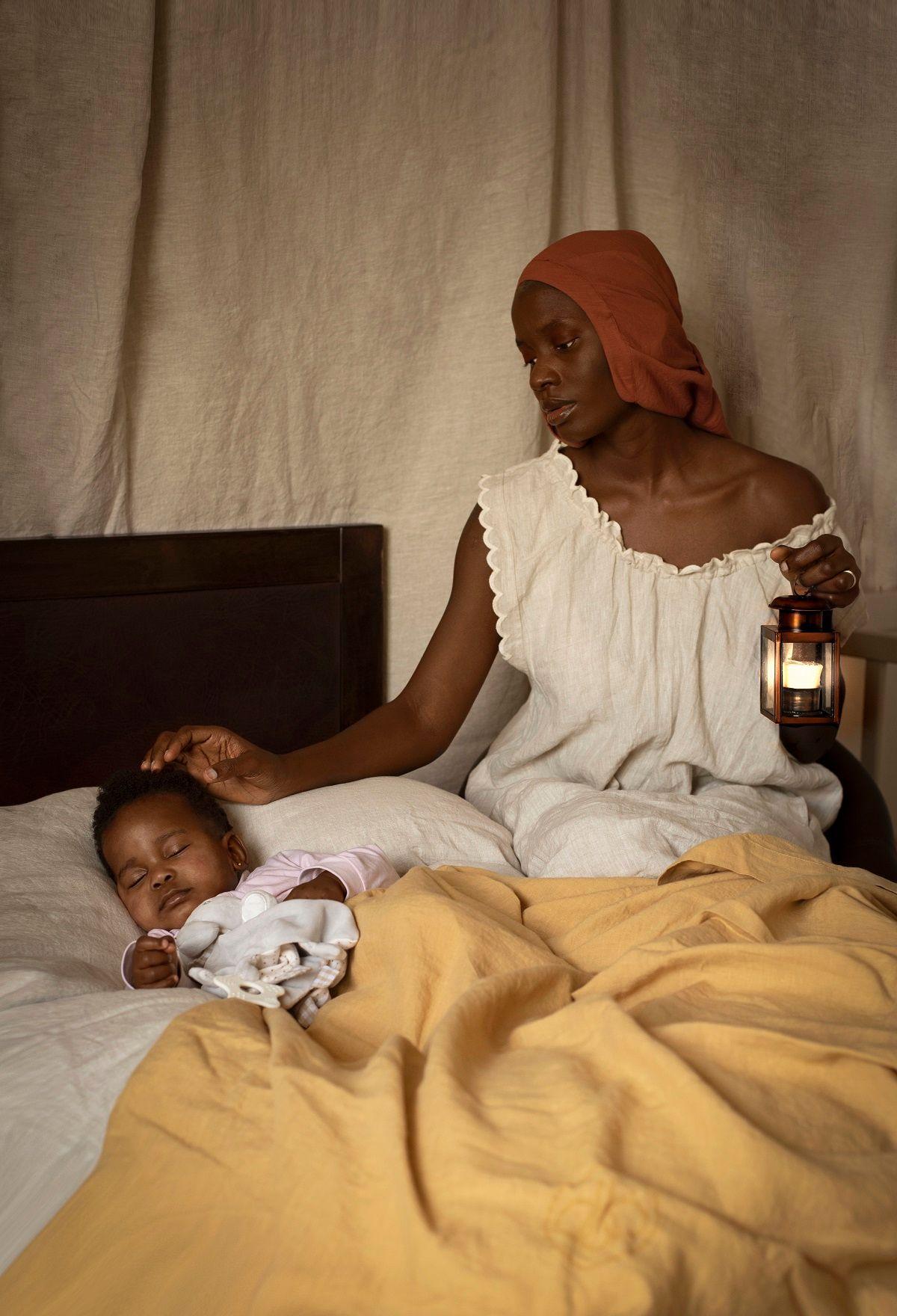 Dola Posh wears an orange headscarf and a white nightie. She is holding a lamp and standing by a bed where her daughter is sleeping.