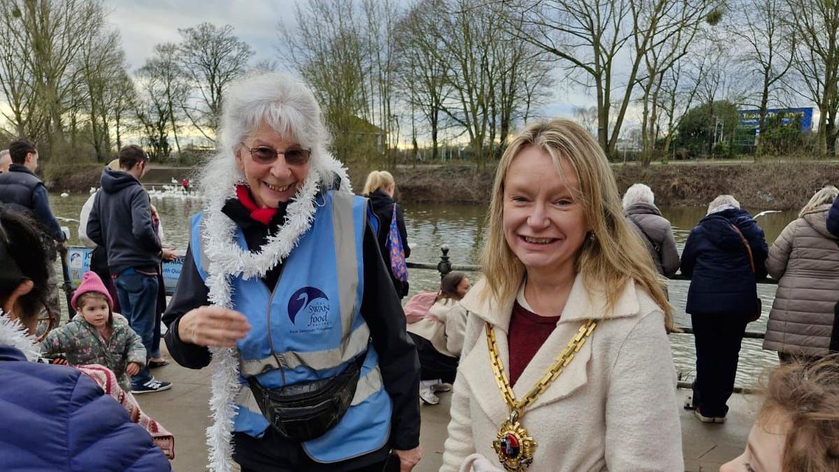 Two women look at the camera among a crowd of people standing along a railing by the river. One of the women has white hair and is wearing a blue Swan Food Project tabard and a string of white tinsel round her neck. The other has long blonde hair, and is wearing a cream coat and mayoral chain. 