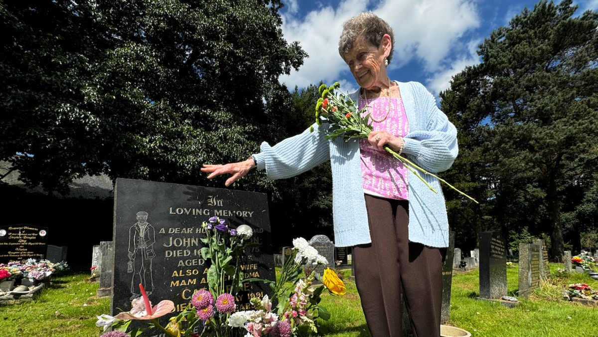 A woman looks at her parents' grave, she is holding flowers and their are trees in the background. The black headstone with gold and black engraving 