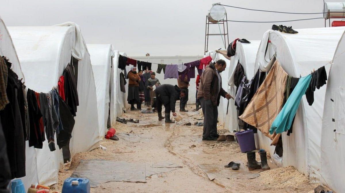 A small group of people stand around looking at two rows of white temporary accommodation tents. The tents have washing hung out to dry in lines stretching between them. The floor is muddy.