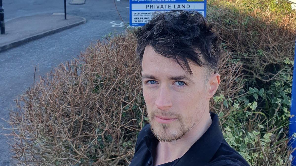 A man in a navy shirt and a beard stands in front of a parking sign