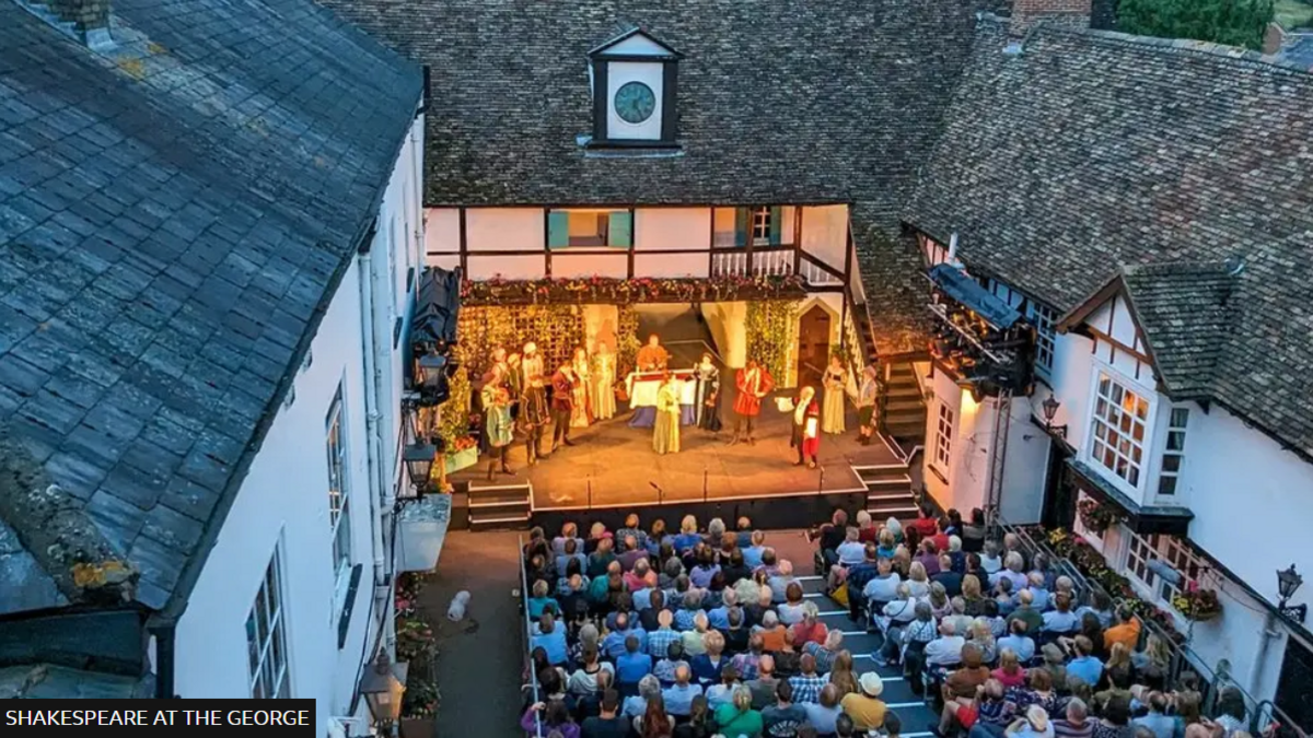 A bird's eye view of a play being performed in the courtyard of an inn. The audience is seated in the open air between two white buildings, while the actors are bathed in yellowish light on a stage in front of them, which is partially open air. There is a clock on the roof above the stage.