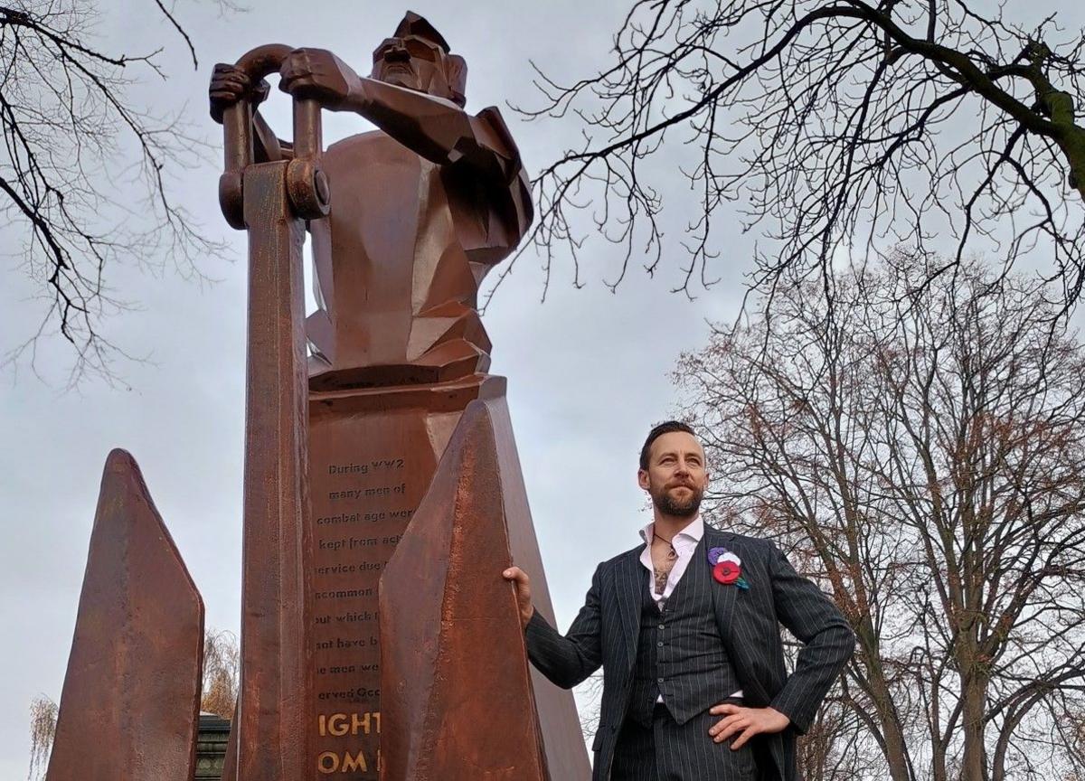 The camera is looking up at the monument, which Luke Perry is standing next to, and there are trees in the background.