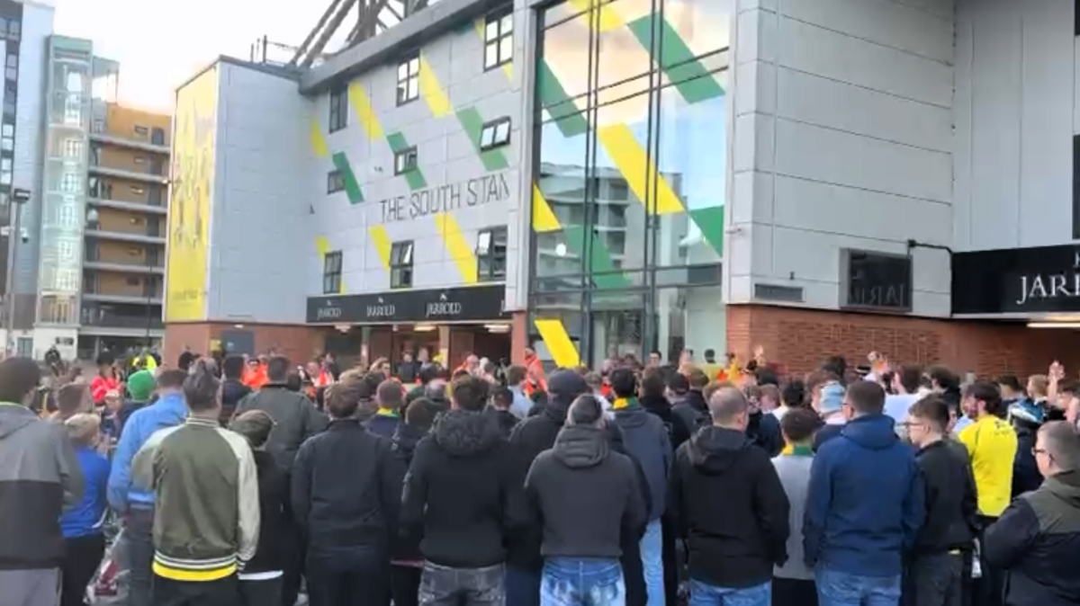 A crowd of around 50 football supporters stand outside the glass doors of an entrance to a football stadium, some with their hands in the air as they chant.