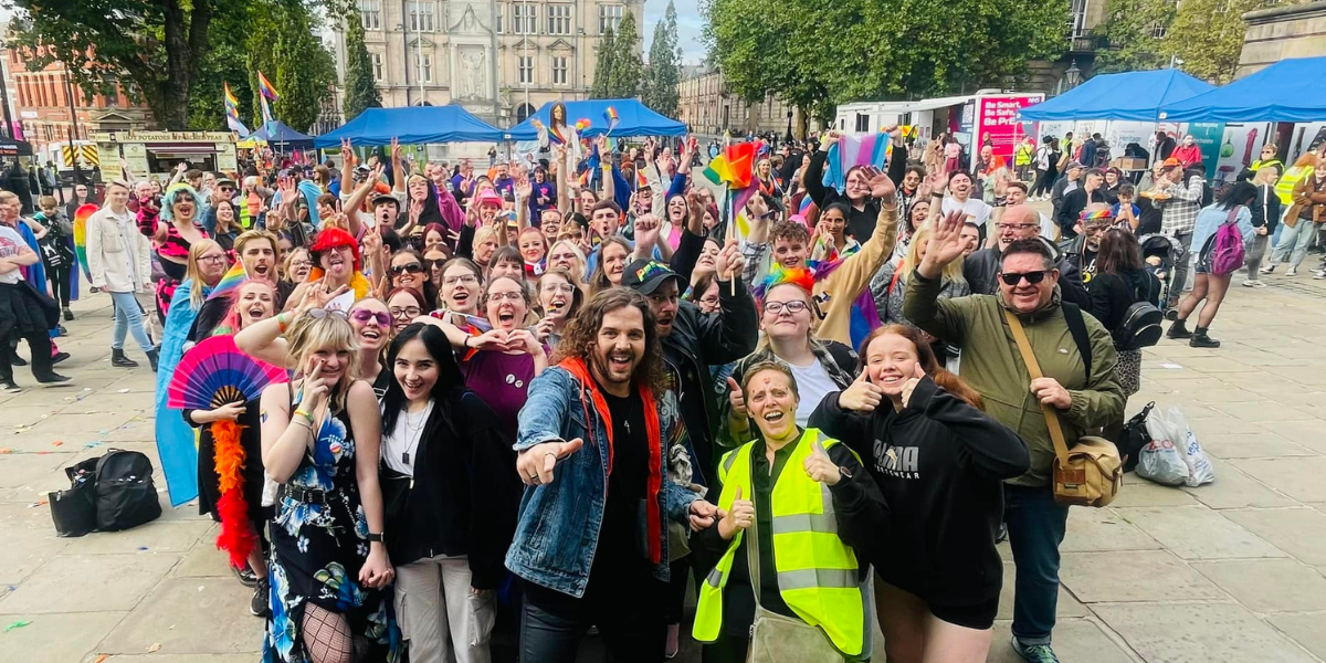 A large crowd of people on Preston's Flag Market, looking and pointing at the camera and smiling, wearing casual clothing and some with rainbow pride flags