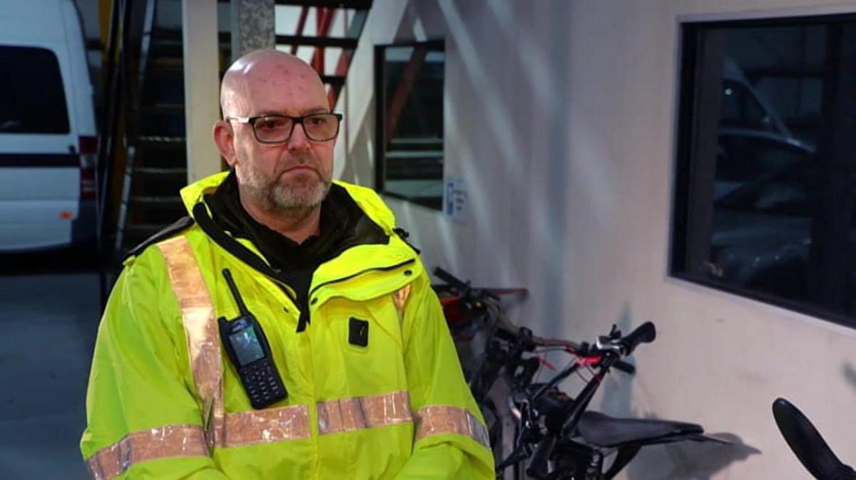 Detective Inspector Tim Evans wearing yellow fluorescent jacket standing in front of a row of bikes. His walkie-talkie is pinned to his chest and he is a bald man with a short white/grey beard and dark rimmed glasses