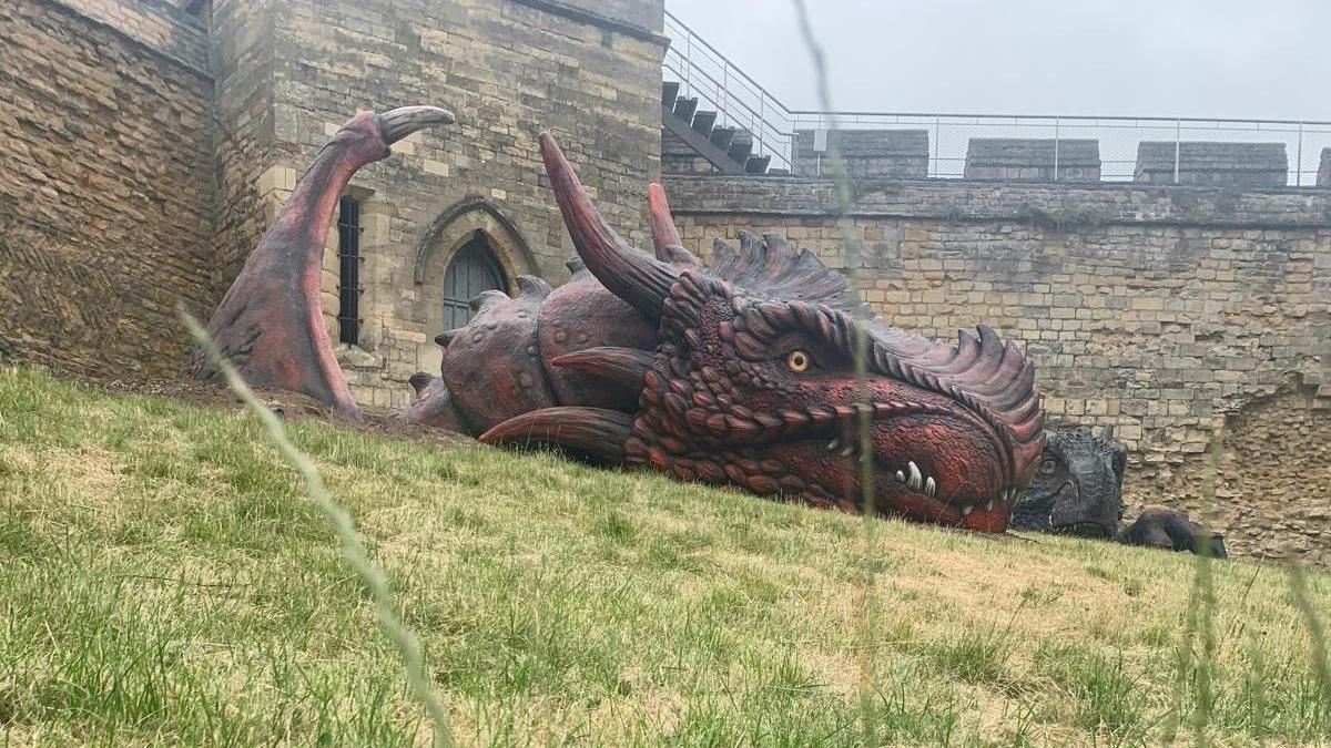 A large dragon sculpture - the dragon has red scales, white teeth and horns. A second dragon sculpture can be seen in the background. Blades of grass can be seen in the foreground.