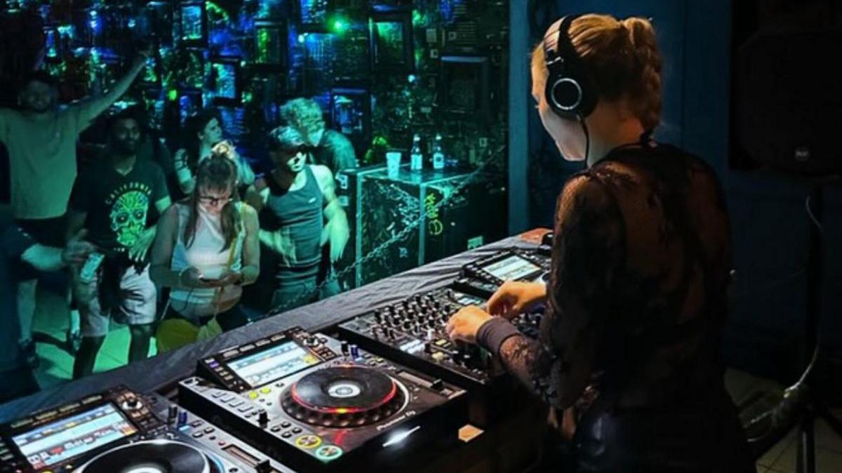A woman wearing a lace black long sleeve top with her hands over a set of DJ decks on stage. In front of her are a couple of people dancing in the crowd under green lighting.
