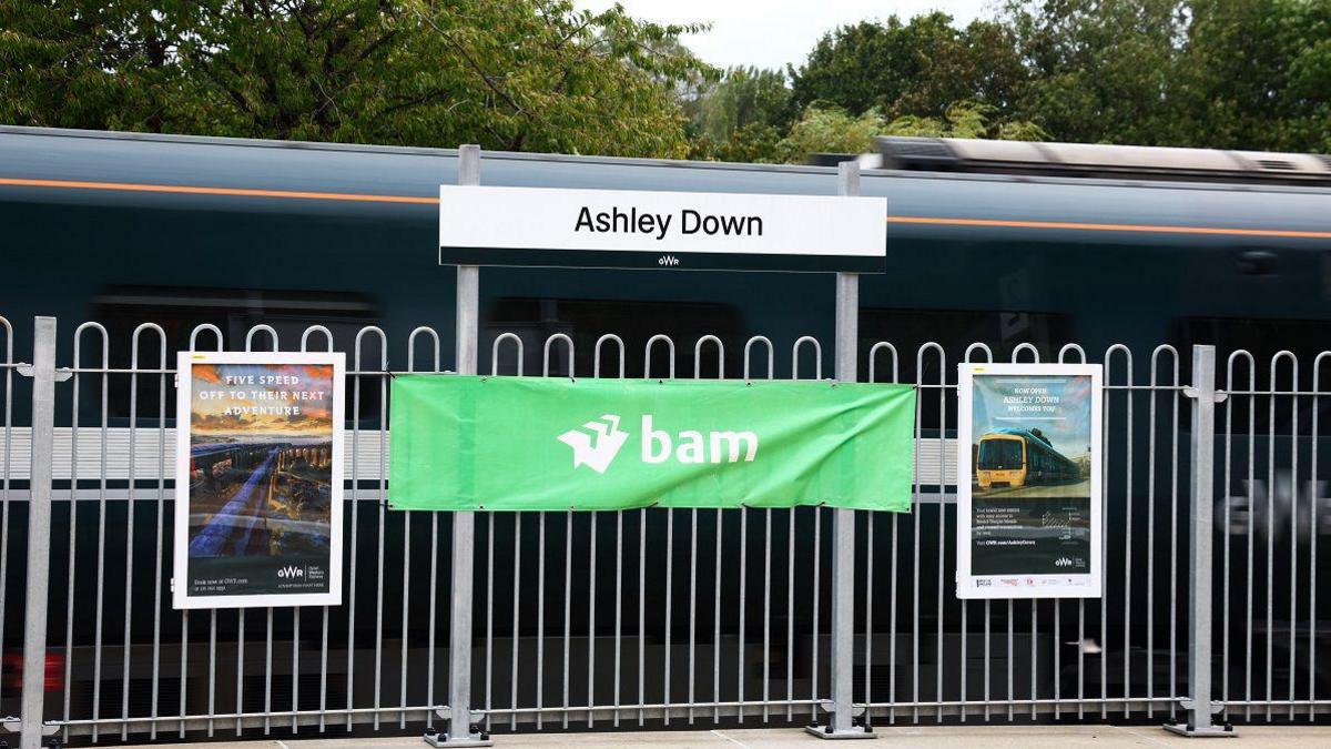 A train platform with a sign which reads 'Ashley Down' and a green Great Western Railway train on the track