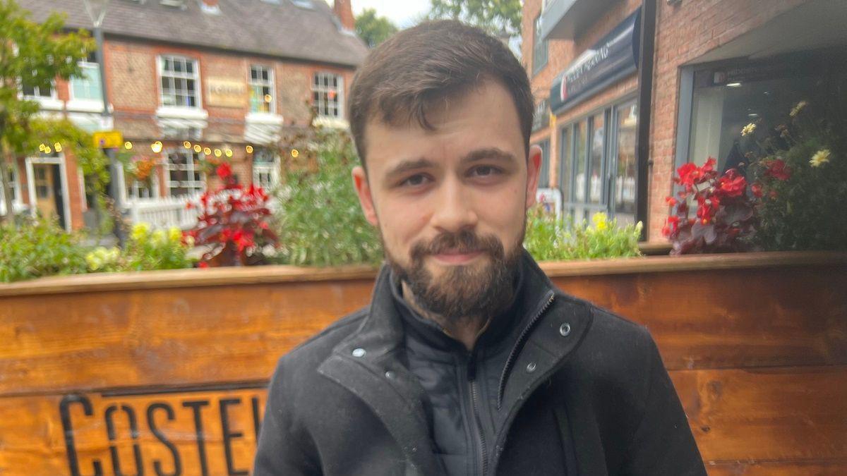 Kenny Caune sitting down at a coffee shop outdoors in Altrincham. He is wearing a dark grey/black winter coat and has a beard. 