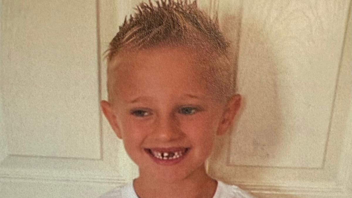 Freddie Farrow with his blond hair spiked up. He is standing in front of a door and smiling while wearing a white T-shirt.