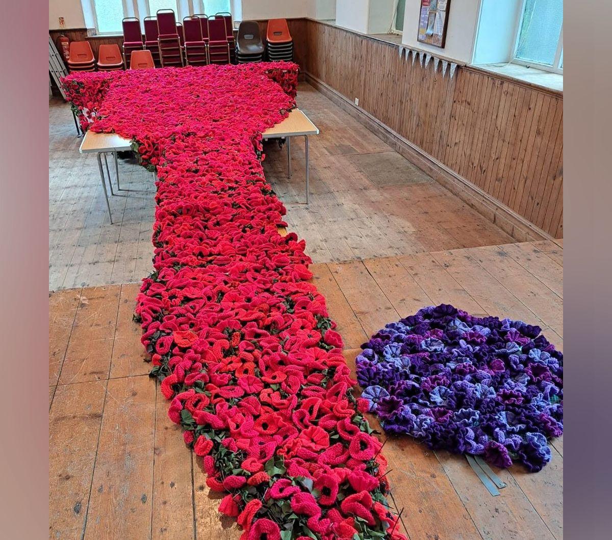 The red poppy display is seen rolled out on a wooden floor. To the side of it is a smaller purple knitwork.