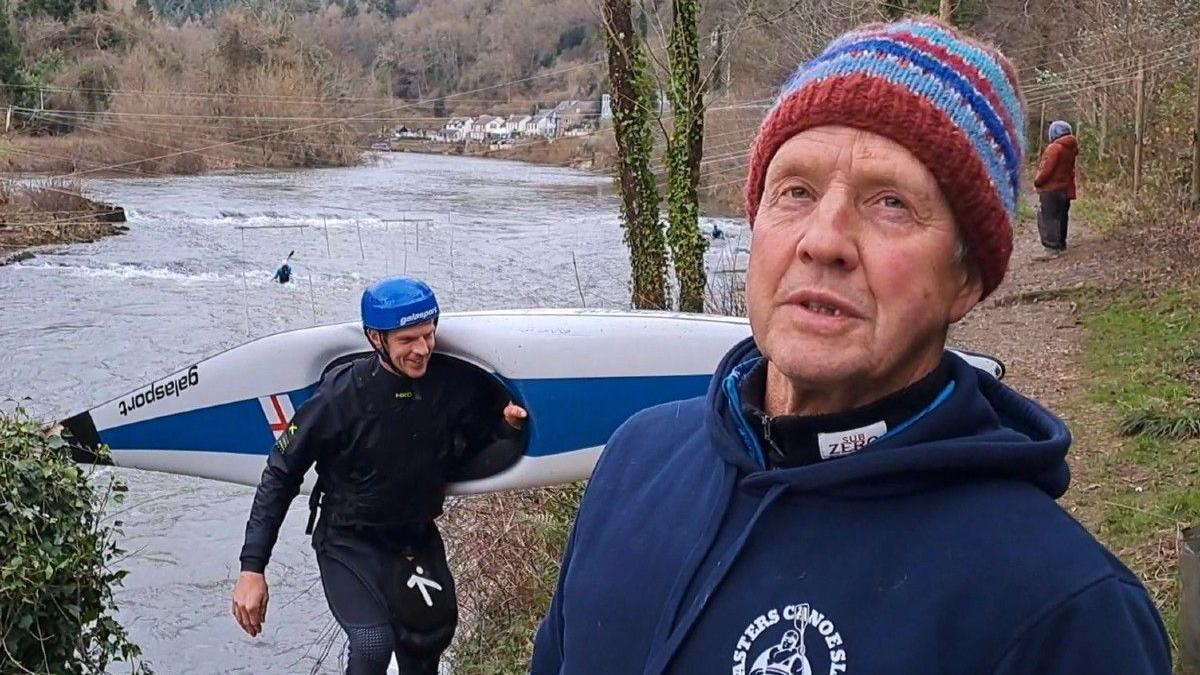 An older man wearing a blue hoodie and a striped wool hat, standing on a river bank. He is talking to the camera. The river is pictured in the background and a man is carrying a canoe over his shoulder, leaving the water. 