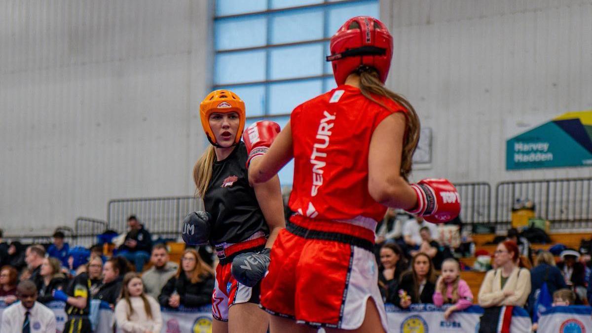 Two women are in a kickboxing tournament. One wears a bright orange sleeveless top and matching shorts, the other is in a black top and shorts. Both are wearing helmets and boxing gloves and are stood up sparring