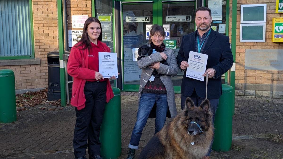 Three people stand outside a building holding certificates in their hands. The person in the middle is holding a small dog and a bigger dog is standing in front of them.