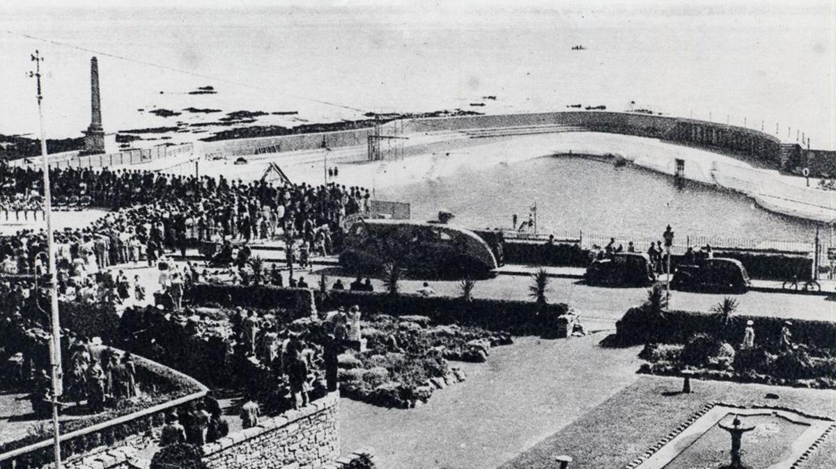 Black and white image of the Jubilee Pool in Cornwall. The image shows hundreds of people standing at the edge of the pool. Cars are parked around the edge.